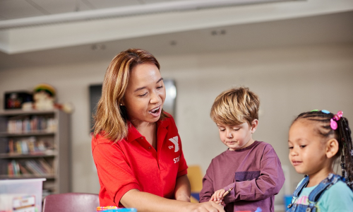 Preschool teacher with a preschool aged boy and girl sitting around a table. The teacher is pointing to the an item on the table,