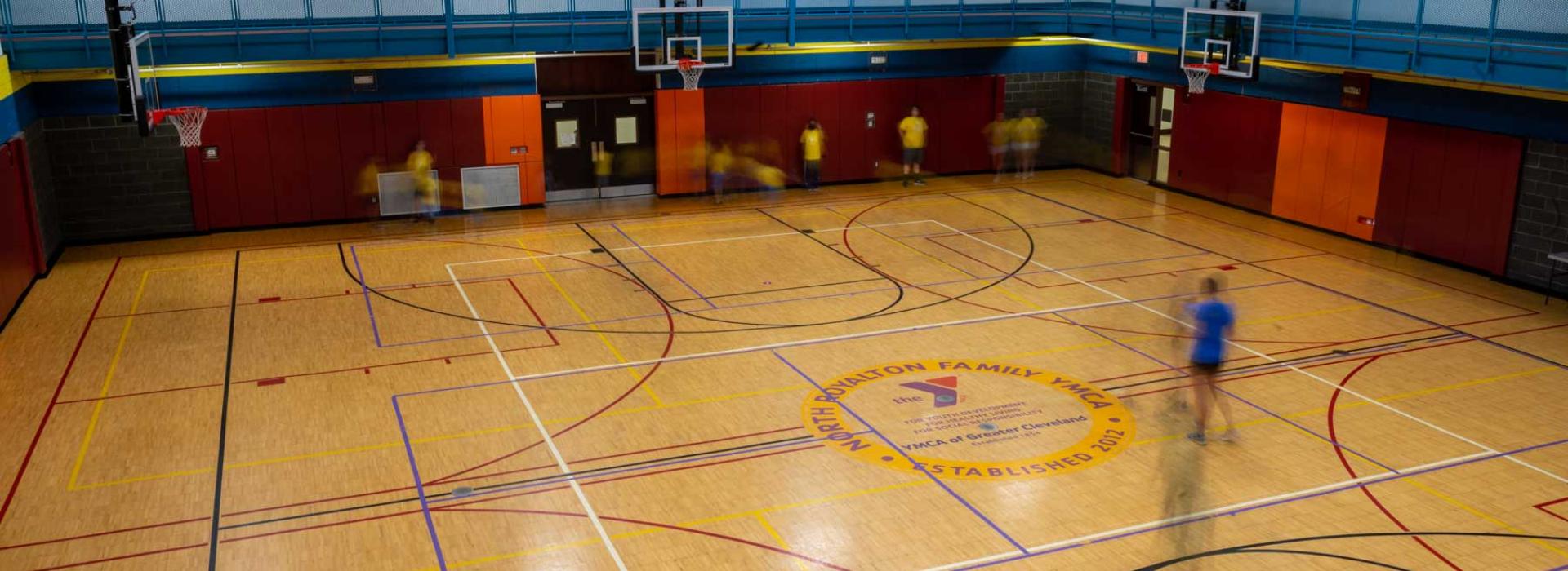 Basketball court marked with lines for multiple sports and split courts. North Royalton YMCA logo in middle of court and members playing basketball.