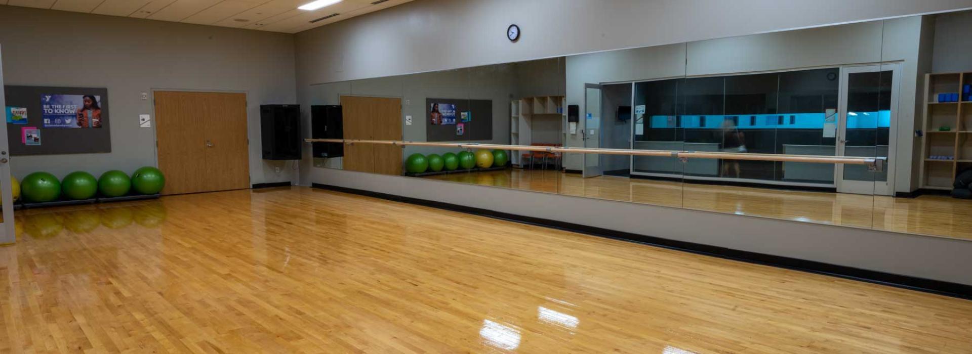 Image shows group fitness room with exercise balls, a mirrored wall with ballet bar, and wood floors.
