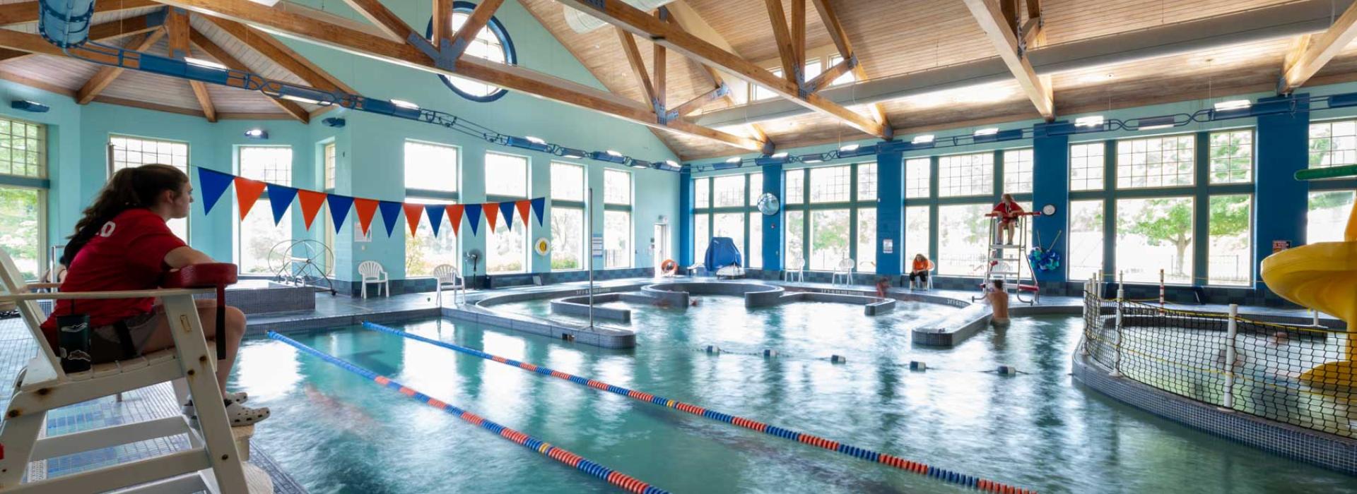 Image shows female lifeguard looking over pool area. Image of pool area includes 3 lanes for swimming, hot tub with "lazy river" around it, kids playing in pool, and jungle-gym water park slide.
