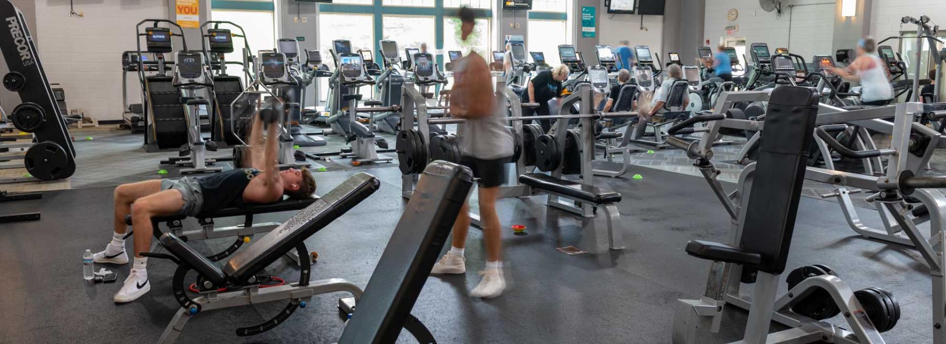 Image shows weight room with members. Pictured: free weights, benches, cardio machines, and stationary bikes.